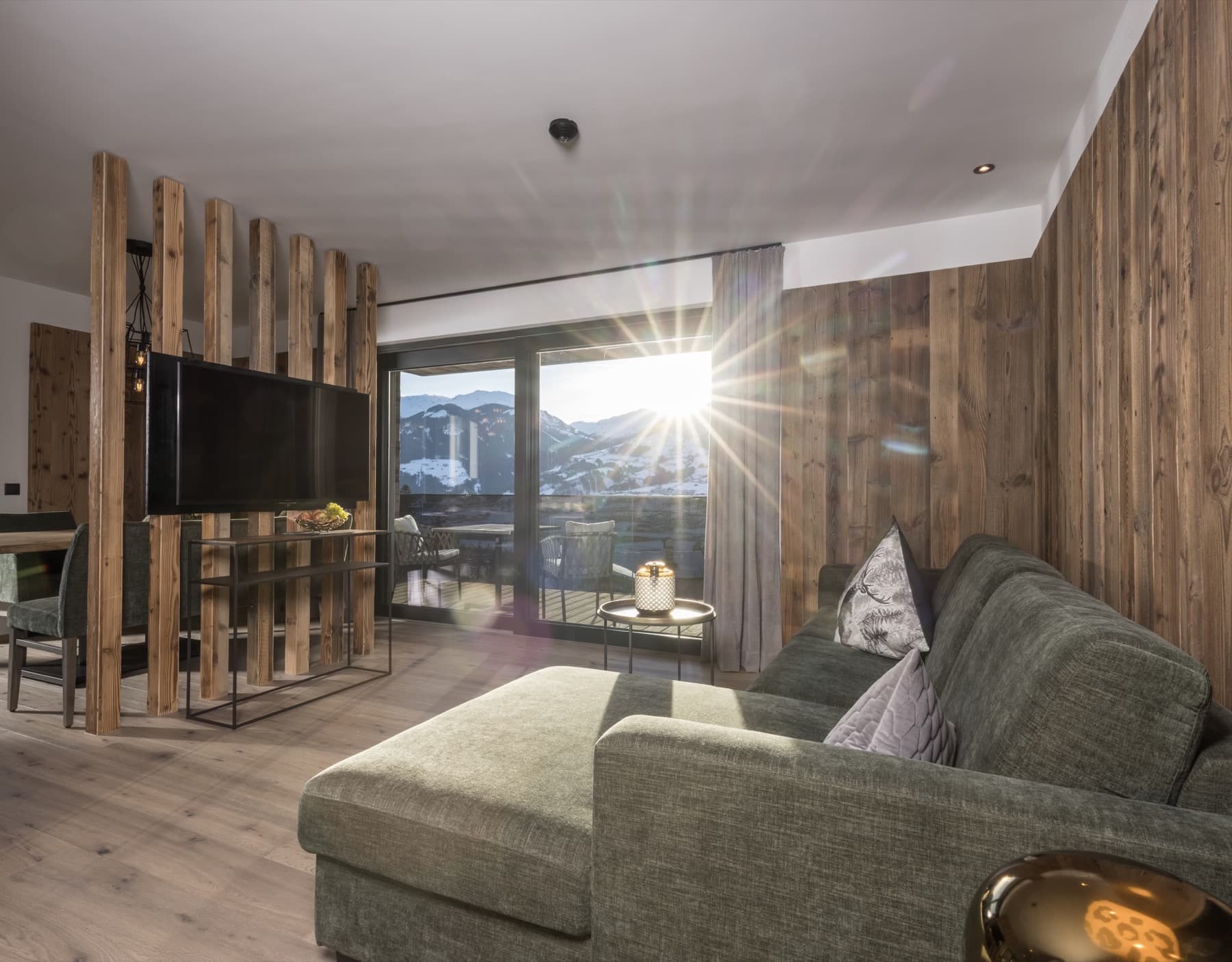 Bright living area with a green corner sofa, TV and rustic wooden walls, looking straight out at the sunny mountains.