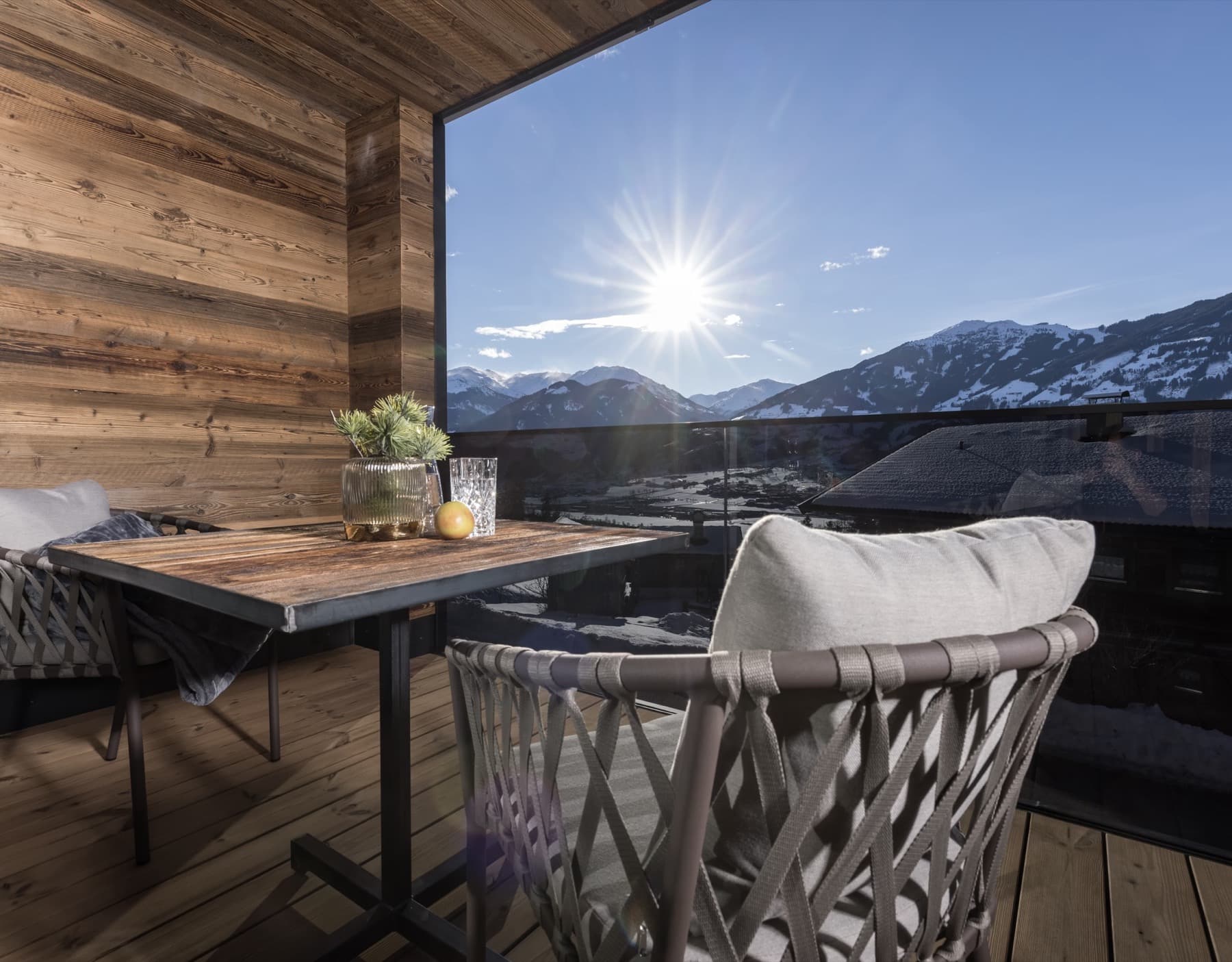 Covered wooden balcony with a table and comfortable seating, looking out on the sunny, snow-covered Zillertal mountains.