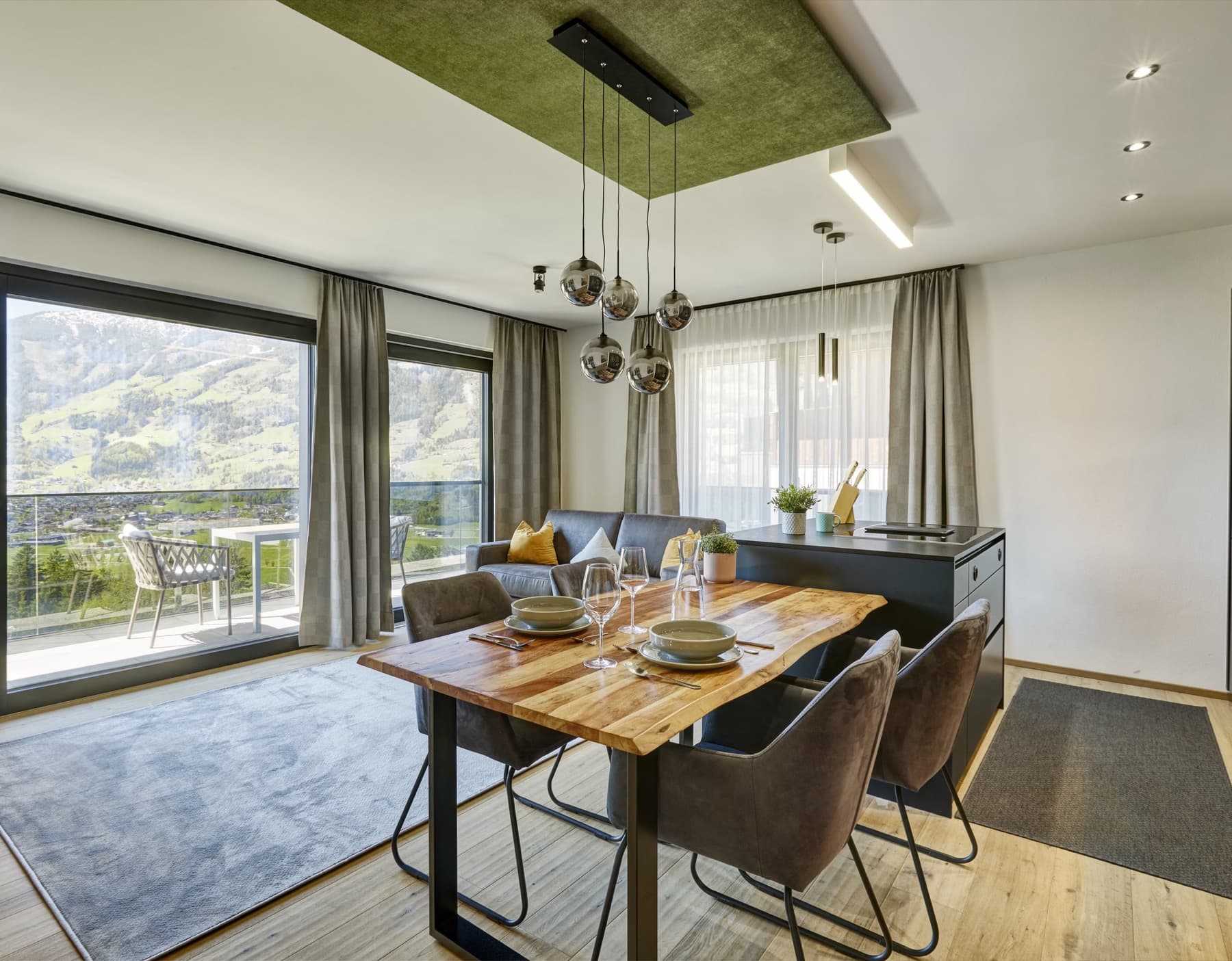 Bright open dining area with a laid wooden table, modern kitchen, cosy sofa and a wide view of the mountains.