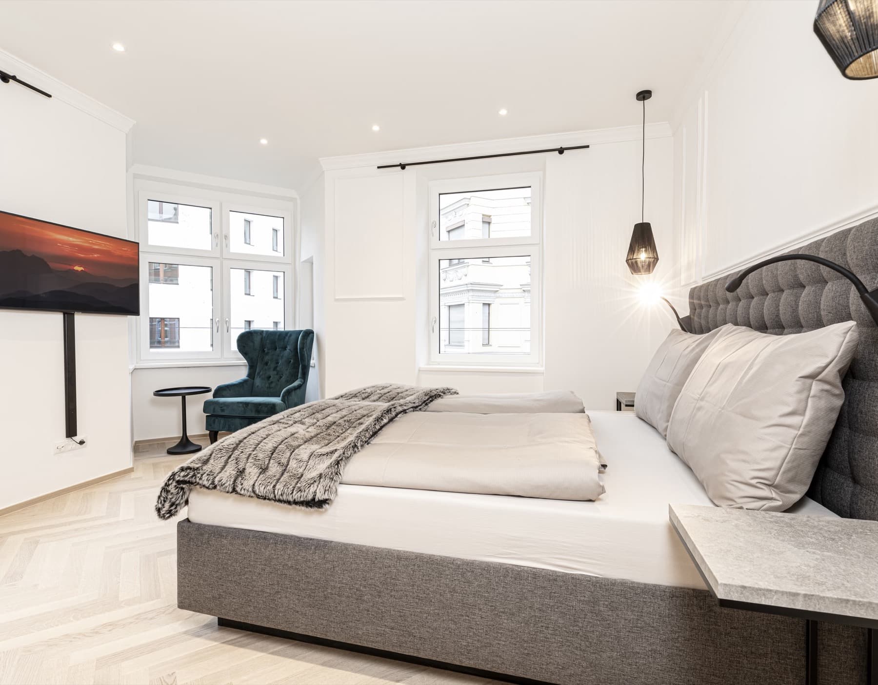 Komfortables Schlafzimmer mit großem Doppelbett, gepolstertem Kopfteil, Fernseher und türkisfarbenem Ohrensessel.