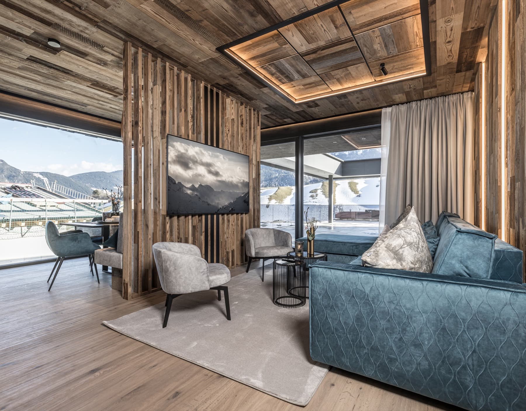 Gemütlicher Wohnbereich mit blauem Sofa, Fernseher an einer Holzlamellenwand und großen Fenstern zur winterlichen Berglandschaft.